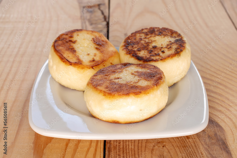 Three homemade cheese pancakes on a white ceramic saucer on a wooden table, close-up.
