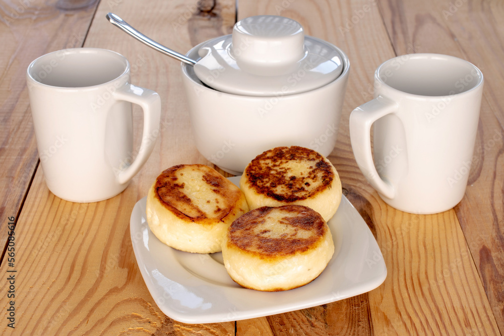 Three homemade cheese pancakes on a white ceramic saucer with white cups on a wooden table, macro. Three homemade cheese pancakes on a white ceramic saucer with white cups on a wooden table, macro.