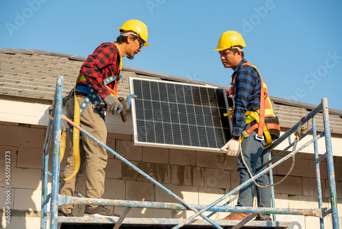 Young technician standing on metal platform installing heavy solar photo voltaic panel on blue sky background. Stand-alone solar panel system installation, efficiency and professionalism concept. © sirisakboakaew