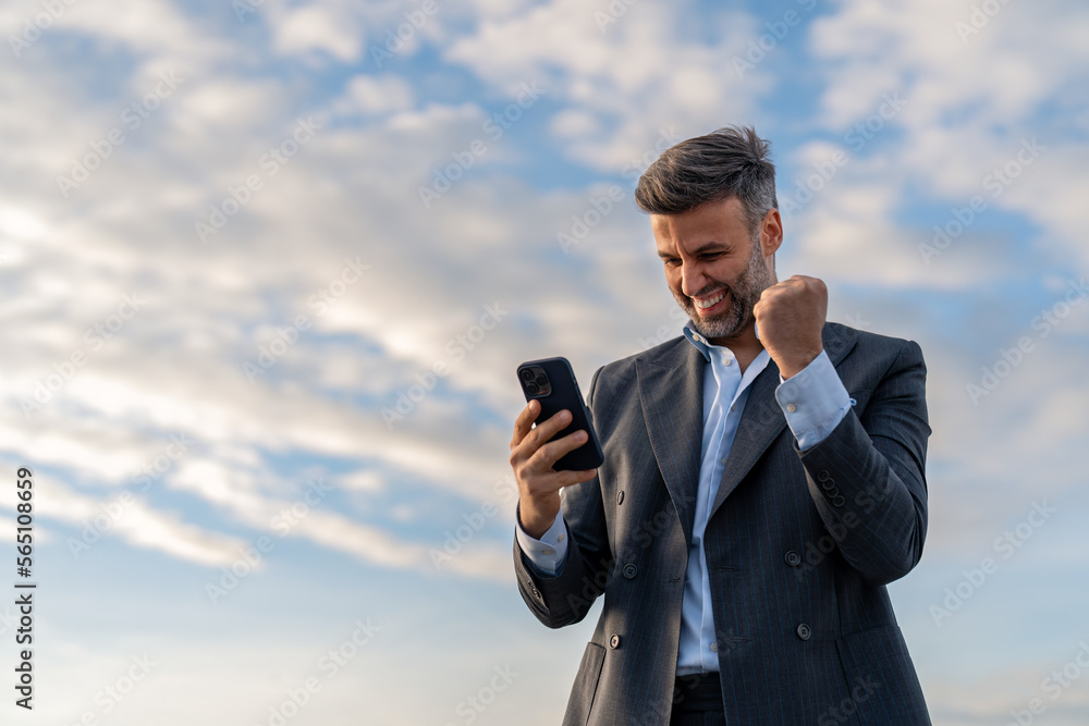Handsome happy businessman feeling excited raising fist in yes gesture looking at cell phone celebrating work success, getting new approved job opportunity succeed in career and financial goals. Handsome happy businessman feeling excited raising fist in yes gesture looking at cell phone celebrating work success, getting new approved job opportunity succeed in career and financial goals.