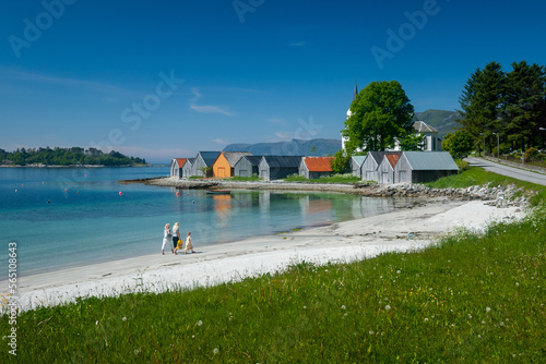 Two individuals stroll along a pristine white beach beside colorful waterfront houses under clear blue skies.