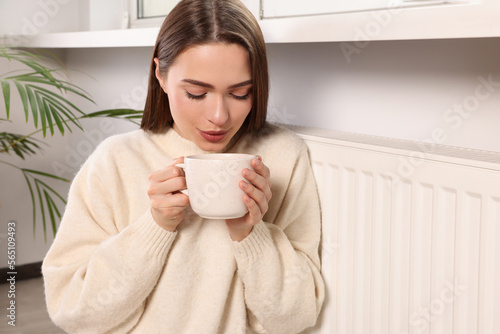 Woman holding cup with hot drink near heating radiator indoors