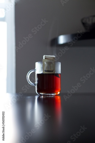 A photograph of a cup of coffee served in a clear glass mug. The coffee is dark in color and appears to be steaming. The mug is resting on a surface and is partially obscured by the steam rising from 