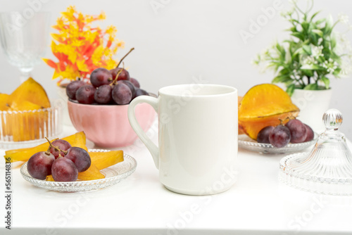 A white blank coffee mug on the top of a white table with the fresh fruits arranged around it