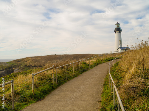 A dirt footpath leading to a beautiful white lighthouse on a high hilly bank overgrown with withered grass. White storm clouds in the sky. Beautiful nature. Romance, tourism, excursions, solitude.