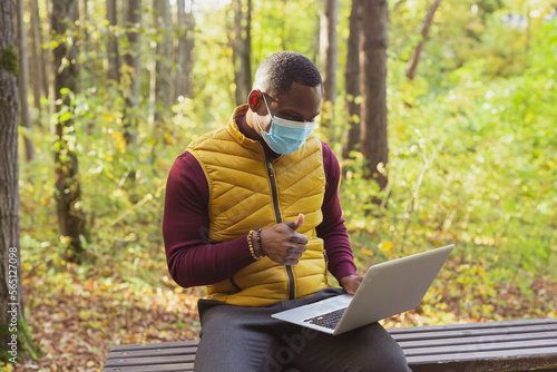 African american man in medical mask sitting in city park on a bench with laptop video call with friend online outdoor. Man freelancer learn working remotely in street e-learning and meeting on