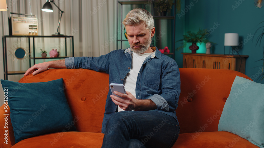 Bearded middle-aged man sitting on sofa uses mobile phone smiles at night home apartment. Mature guy texting share messages content on smartphone social media applications online, watching relax movie Bearded middle-aged man sitting on sofa uses mobile phone smiles at night home apartment. Mature guy texting share messages content on smartphone social media applications online, watching relax movie