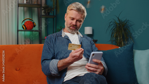 Middle-aged old man using credit bank card and smartphone while transferring money, purchases online shopping, order food delivery at home apartment indoors. Mature senior guy in evening room on couch