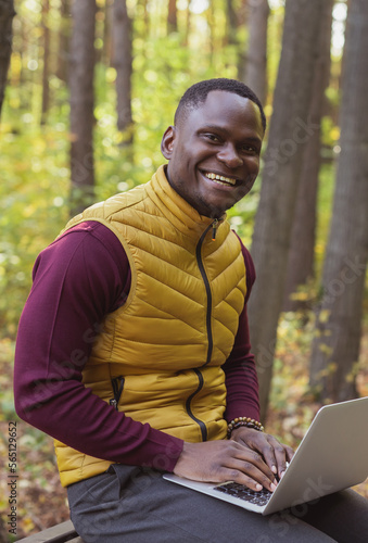 Portrait African american man sitting in city park on a bench with laptop studying online outdoors. Man freelancer learn working remotely in street e-learning vertical banner