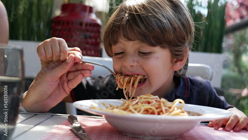 Child sitting at lunch table eating spaghetti. Mother hand feeding pasta to son at restaurant. Noodles with red sauce on plate