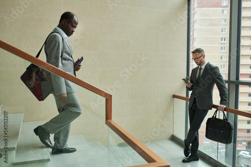 Mature African American businessman walking downstairs while young male entrepreneur using smartphone inside modern business center