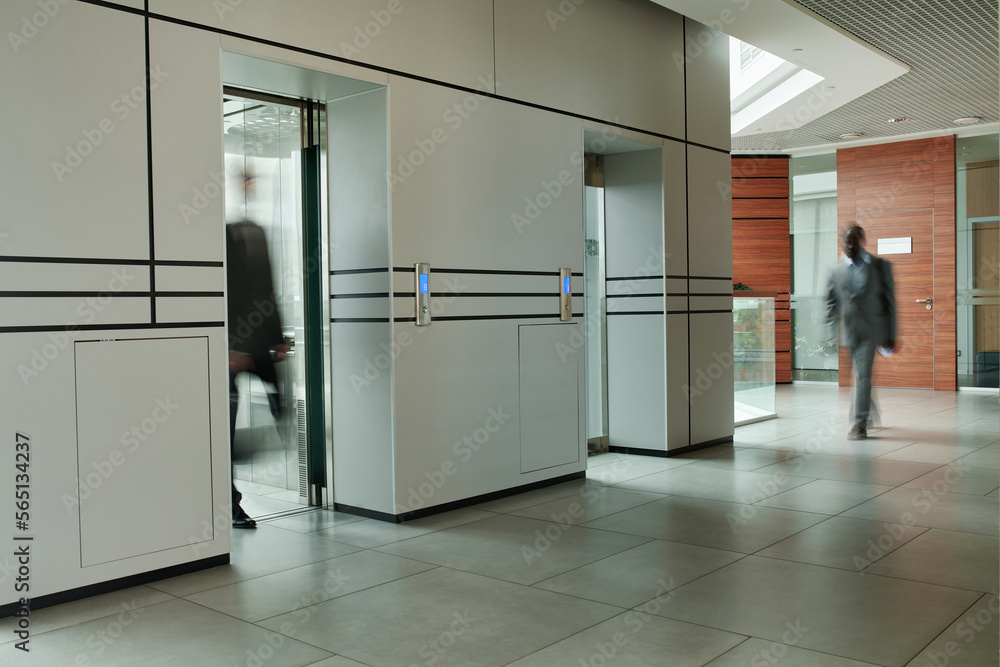 Blurry motion of two male employees in spacious corridor of modern business center while one of them getting out of elevator Blurry motion of two male employees in spacious corridor of modern business center while one of them getting out of elevator