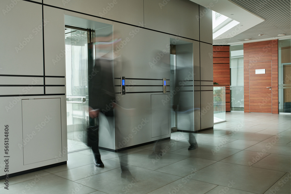 Part of spacious corridor inside modern office center and two blurred businessmen by elevators with glass doors moving along