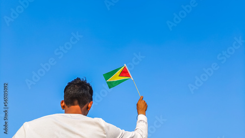 Boy holding Guyana flag against clear blue sky. Man hand waving Guyanese flag view from back, copy space