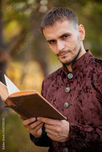 A young man in medieval brown clothes stands with an old book in his hand. A guy in a historical costume in nature.