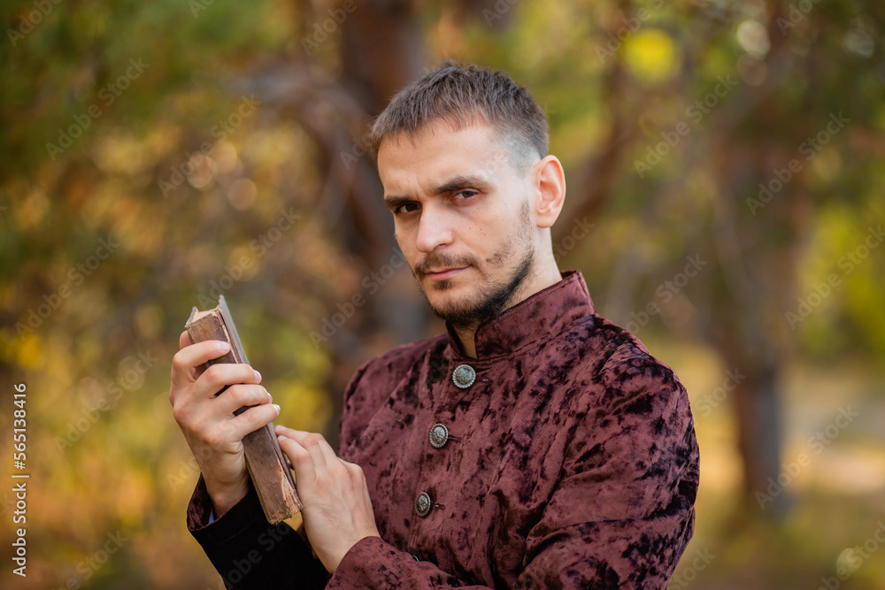 A young man in medieval brown clothes stands with an old book in his hand. A guy in a historical costume in nature. A young man in medieval brown clothes stands with an old book in his hand. A guy in a historical costume in nature.