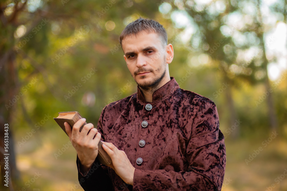 A young man in medieval brown clothes stands with an old book in his hand. A guy in a historical costume in nature. A young man in medieval brown clothes stands with an old book in his hand. A guy in a historical costume in nature.