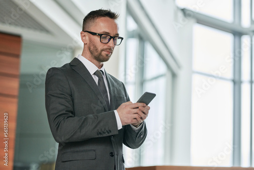 Young serious male broker in formalwear texting in mobile phone while communicating with colleague or client inside office center