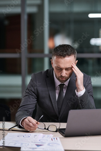 Overworked and tired businessman in formalwear sitting by desk, bending over laptop and touching temple while trying to concentrate