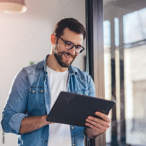 Smiling young businessman working online with digital tablet while standing by window.