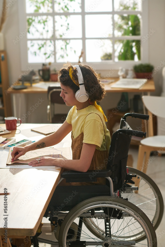 Side view of young creative woman in wheelchair drawing with crayons on paper and listening to music in headphones in home studio Side view of young creative woman in wheelchair drawing with crayons on paper and listening to music in headphones in home studio