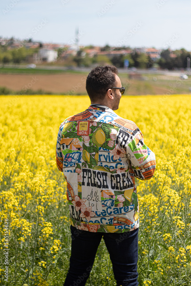 Man in Canola Field. He is wearing jacket with writings and pictures on it. 