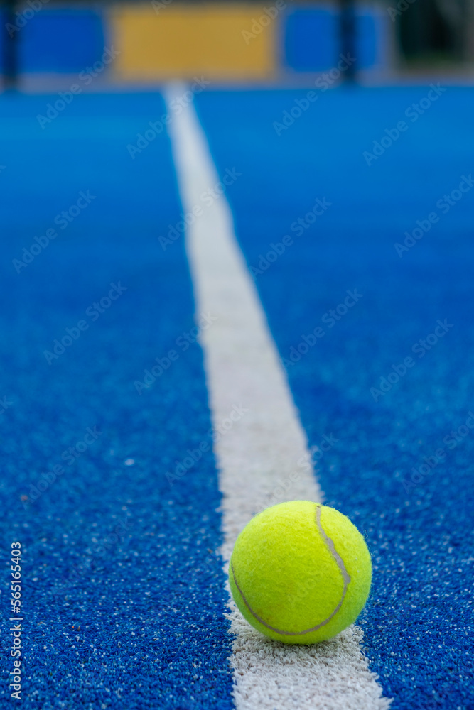 A paddle tennis ball in the foreground on the line of a blue paddle tennis court, racket sports concept
