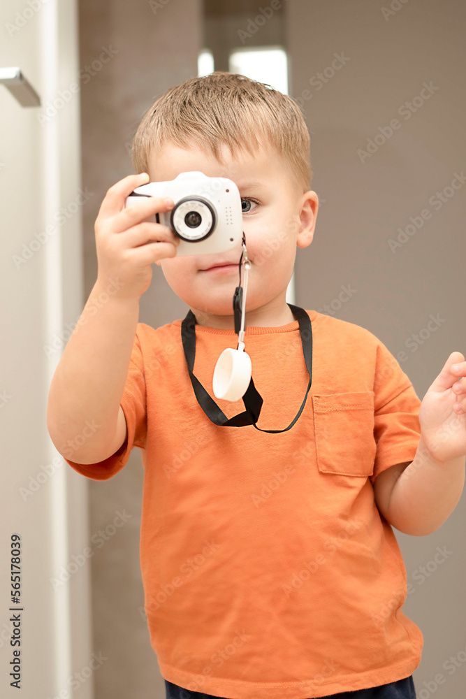 Fototapeta premium People. Handsome funny and emotional caucasian blond little boy in an orange t-shirt with a children's white camera takes pictures in the home interior.