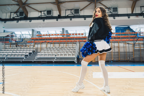 Horizontal shot of cheerleader looking over the shoulder posing with blue shiny pom-poms on basketball court. High quality photo photo