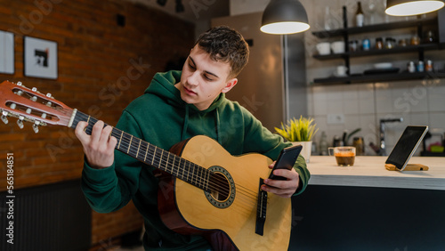 One young man Caucasian teenager sit at home in room playing guitar use smartphone mobile phone to learn for online lesson or course tutorial copy space
