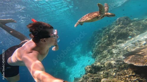 A brown-haired man in a red snorkel mask swims underwater next to a sea turtle and coral reefs. The man spends his best weekend in the Bahamas diving underwater and exploring seabed. Turtle and diver.