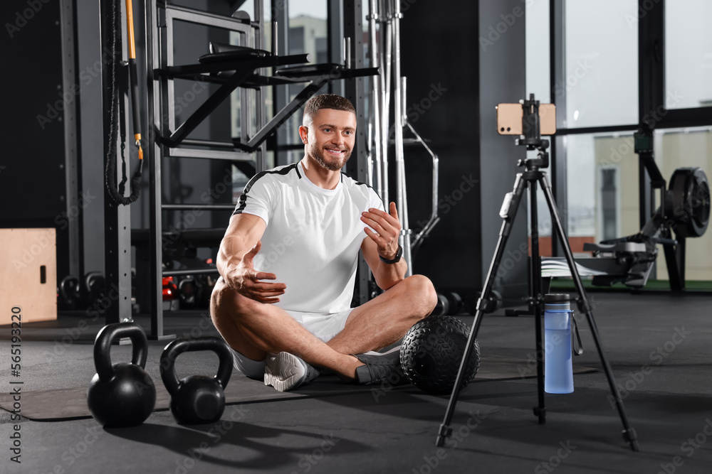Fototapeta premium Man with ball and dumbbells streaming online training on phone at gym. Fitness coach