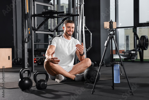 Man with ball and dumbbells streaming online training on phone at gym. Fitness coach