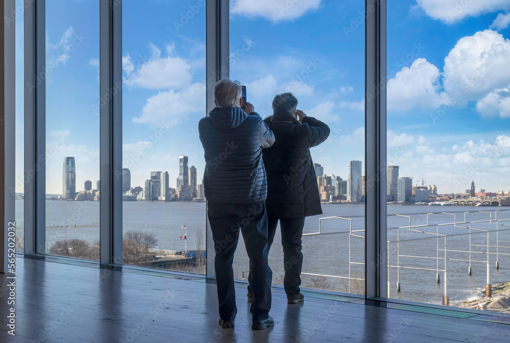 Two men dressed in winter clothes taking pictures of the Jersey City skyline with their phones through large windows, daytime, blue skies and clouds Two men dressed in winter clothes taking pictures of the Jersey City skyline with their phones through large windows, daytime, blue skies and clouds