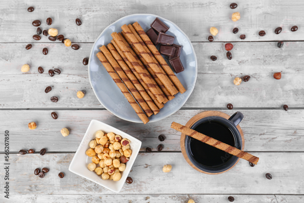 Plate with delicious rolled wafer cookies, cup of coffee and nuts on light wooden background Plate with delicious rolled wafer cookies, cup of coffee and nuts on light wooden background