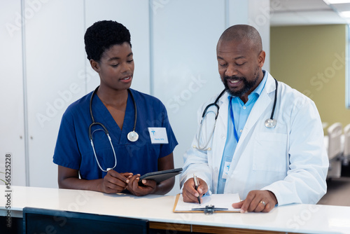African american male and female doctor talking and writing notes in hospital corridor