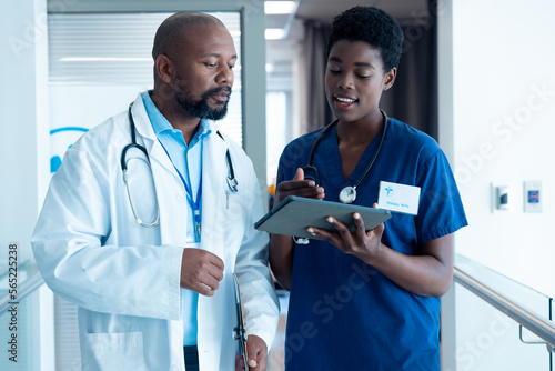 African american male and female doctor using tablet, talking in hospital corridor with copy space