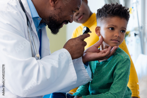 African american male doctor using otoscope to examine ear of boy patient, with mother
