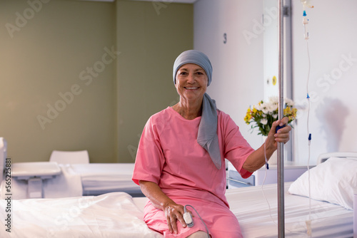 Happy senior caucasian female cancer patient wearing head scarf sitting on bed at hospital photo