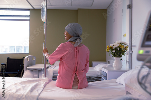 Senior caucasian female cancer patient wearing head scarf sitting on bed with drip at hospital