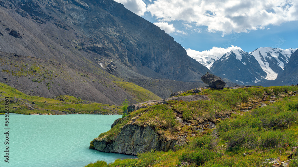 Turquoise mountain lake Karakabak against the background of peaks with snow of glaciers during the day flows down from above in Altai in summer near green grass and hanging stone.