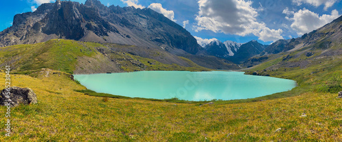 Panorama of the turquoise mountain lake Karakabak against the background of peaks and stone rocks with glaciers and snow with a day in Altai in the summer among the green grass.
