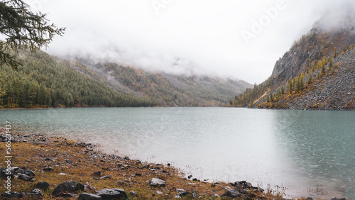 The rain falls on the clear mountain turquoise lake Shavlinskoye with tents on the shore among the rocks in the fogs in Altai during the day. photo