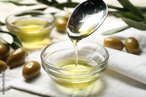 Spoon with cooking oil over bowl and olives on white wooden table, closeup