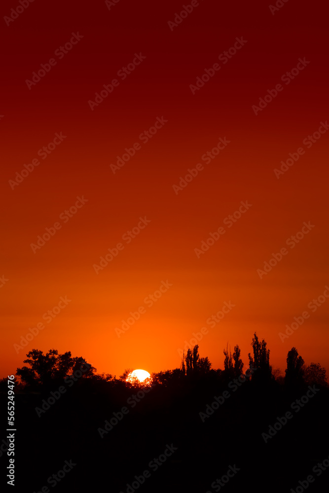Sunset over a lake with ray cast on the lake, treeline in silhouette in the background during summer and sunburst. Big round sun is rising behind the trees over the water, orange and red dramatic sky Sunset over a lake with ray cast on the lake, treeline in silhouette in the background during summer and sunburst. Big round sun is rising behind the trees over the water, orange and red dramatic sky