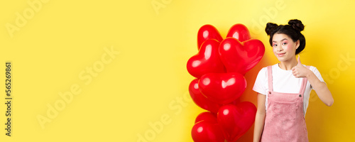 Fashionble asian woman in romantic outfit with make-up, showing thumb up and smiling, praising Valentines day offer, standing near red heart balloons, yellow background photo