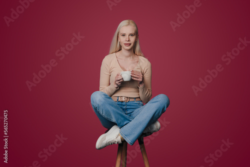 Full length body size photo of woman holding cup of cappuccino sitting on chair and looking at camera isolated on crimson color background.