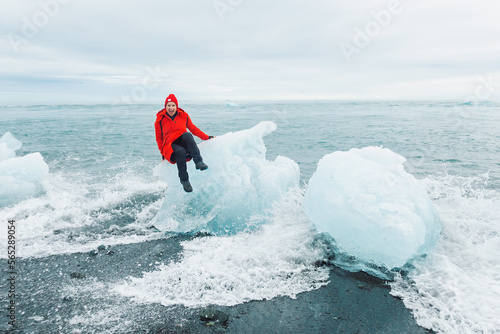 A cheerful tourist in a red jacket sits on a large piece of ice at Jokulsarlon glacial lagoon near Vatnajokull National Park, southeast Iceland photo