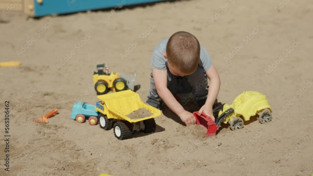 little kid boy playing toy track car on sand children playground in summer sunny day outdoors. male child sitting on ground play toy excavator pretending building site. child digs sand bucket elevator little kid boy playing toy track car on sand children playground in summer sunny day outdoors. male child sitting on ground play toy excavator pretending building site. child digs sand bucket elevator