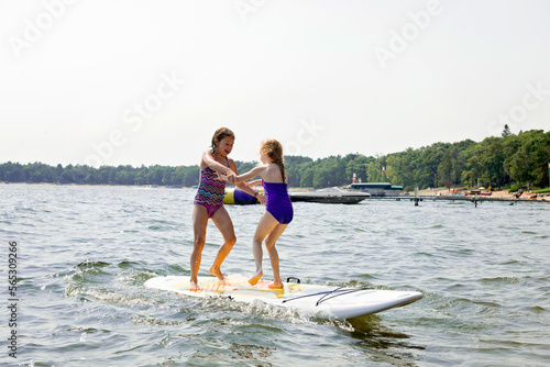 Full length of cheerful sisters standing on paddleboard in lake against clear sky during sunny day photo
